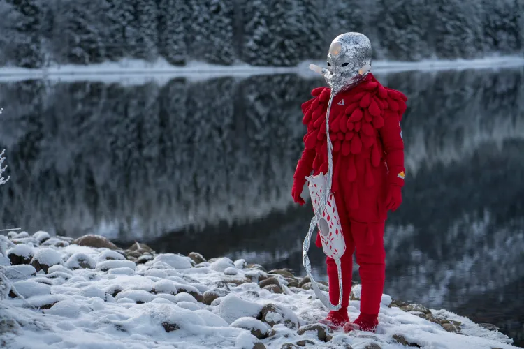 En person i rød teksturert drakt og sølvmaske står på snødekte steiner ved en rolig innsjø med speilvendte furutrær